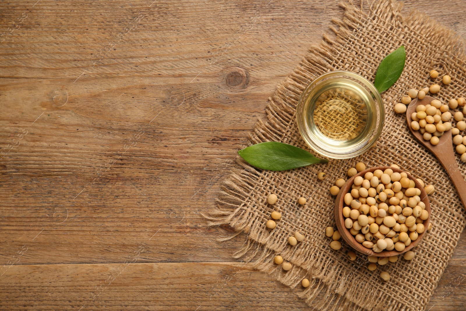 Photo of Glass bowl with oil, leaves and soybeans on wooden table, flat lay. Space for text