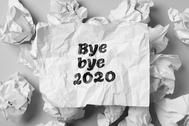 Photo of Crumpled sheet with phrase Bye Bye 2020 among paper balls on light grey table, flat lay