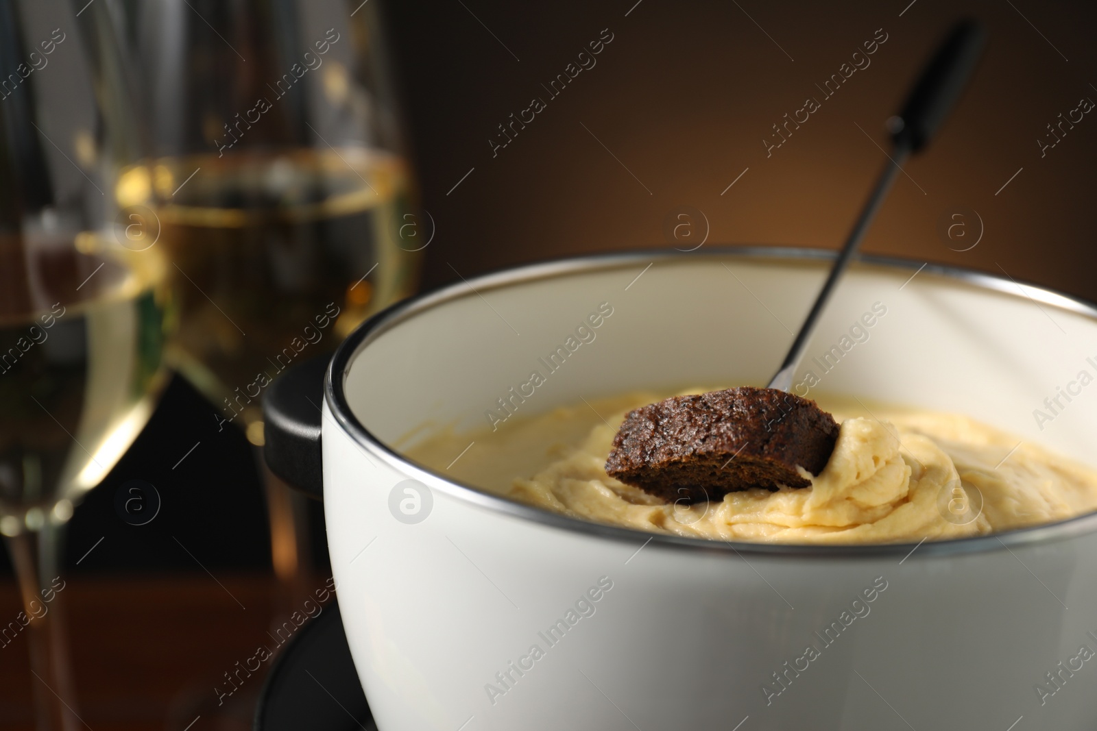 Photo of Fork with piece of bread and melted cheese in fondue pot on dark background, closeup