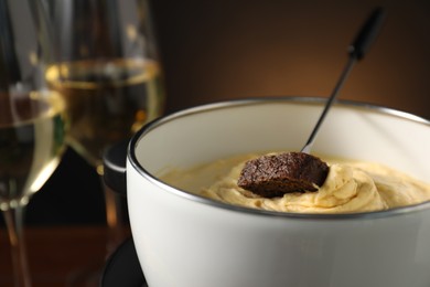 Fork with piece of bread and melted cheese in fondue pot on dark background, closeup