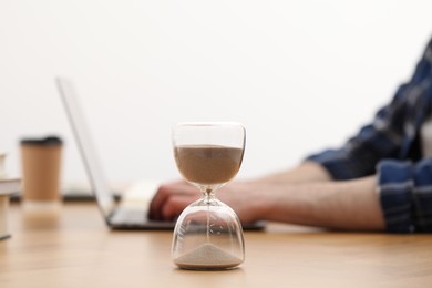 Photo of Hourglass with flowing sand on desk. Man using laptop indoors, selective focus