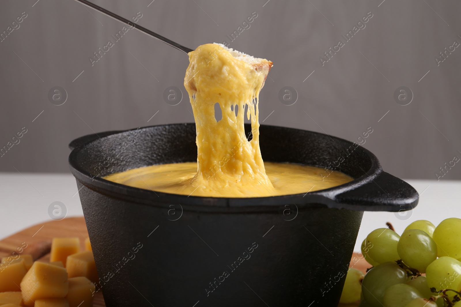 Photo of Dipping piece of bread into fondue pot with tasty melted cheese at table against gray background, closeup