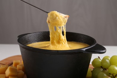 Dipping piece of bread into fondue pot with tasty melted cheese at table against gray background, closeup