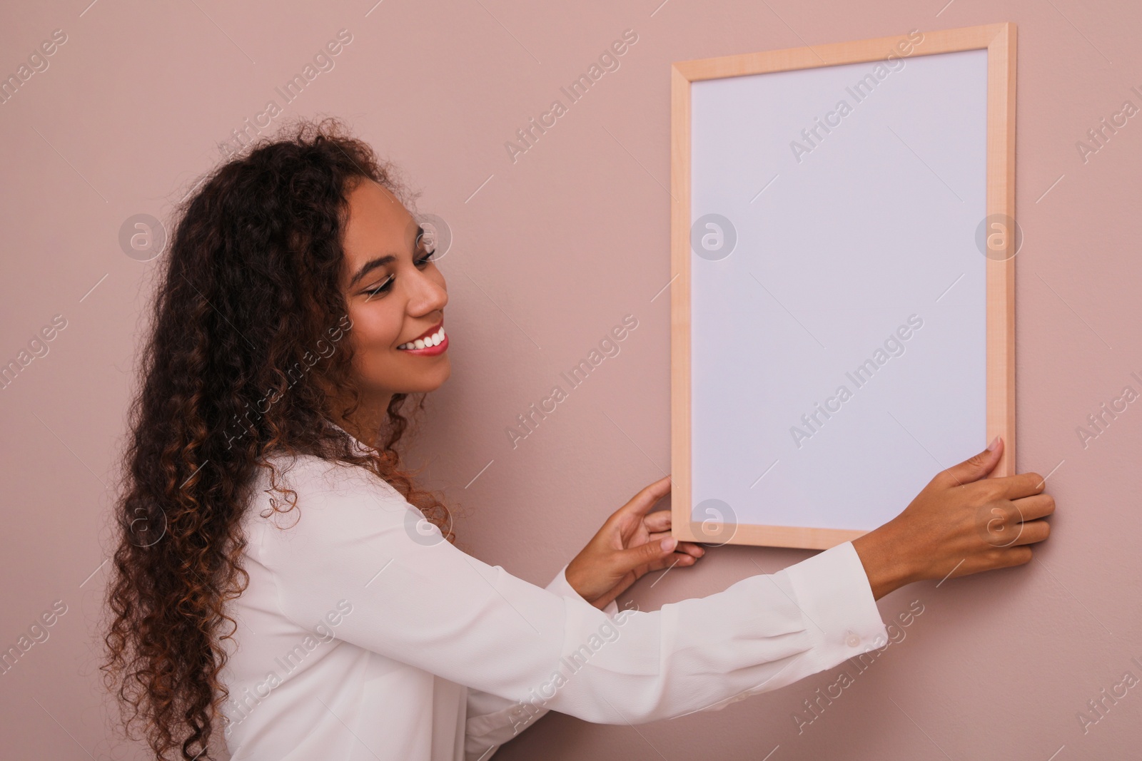 Photo of Happy African American woman hanging empty frame on pale rose wall. Mockup for design