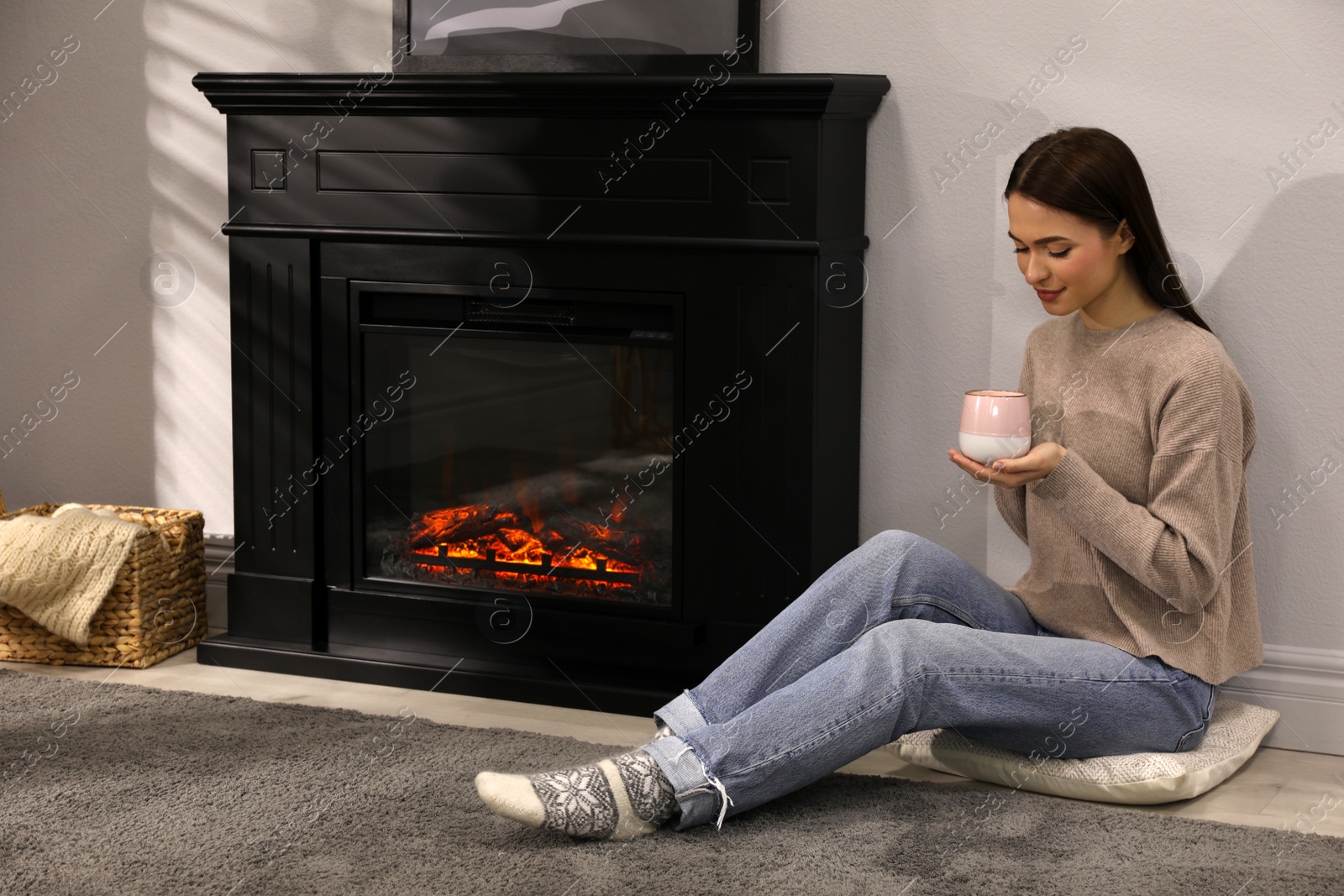 Photo of Beautiful young woman with cup of hot drink sitting on floor near fireplace at home. Space for text