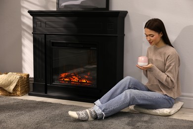 Photo of Beautiful young woman with cup of hot drink sitting on floor near fireplace at home. Space for text
