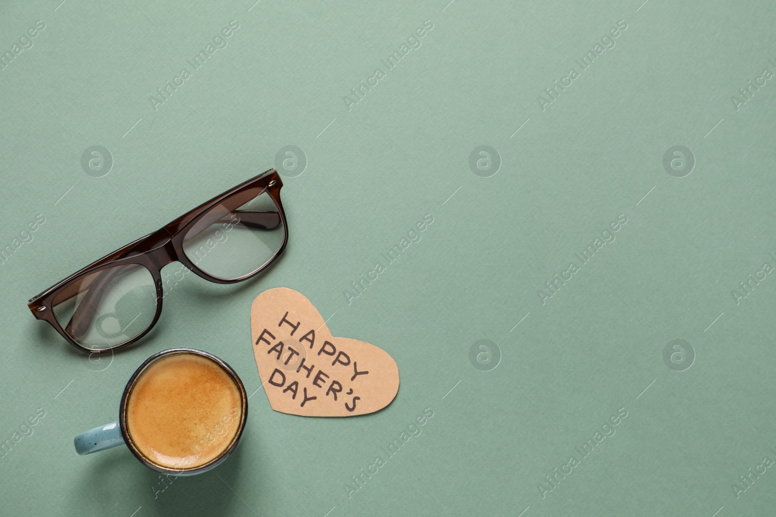 Photo of Heart shaped card with phrase Happy Father's Day, glasses and cup of aromatic coffee on green background, flat lay. Space for text