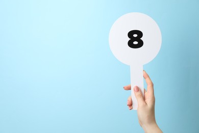 Photo of Woman holding auction paddle with number 8 on light blue background, closeup. Space for text