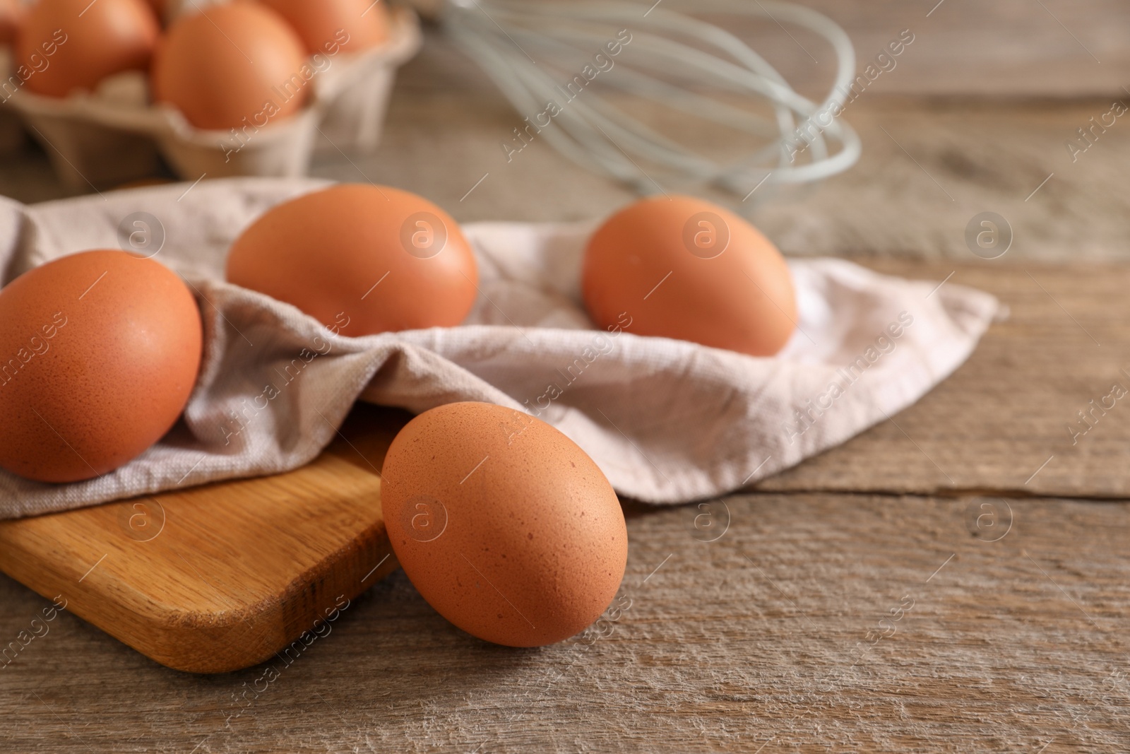 Photo of Raw chicken eggs, napkin, board and whisk on wooden table, closeup