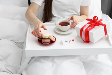 Photo of Tasty breakfast served in bed. Woman with tea, macarons, gift box and I Love You card at home, closeup