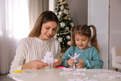 Photo of Happy mother and daughter making paper snowflakes at table near Christmas tree indoors