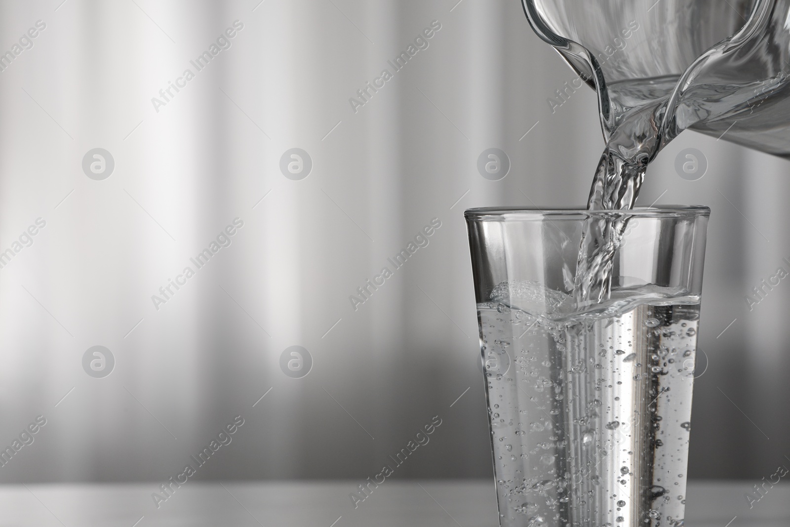 Photo of Pouring water from jug into glass on blurred background, closeup. Space for text