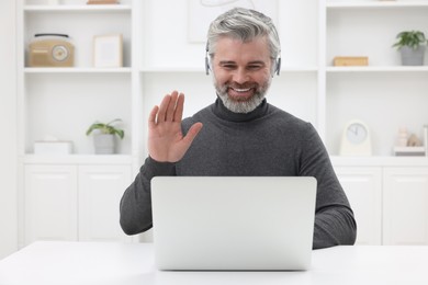 Photo of Man in headphones waving hello during video chat via laptop at home