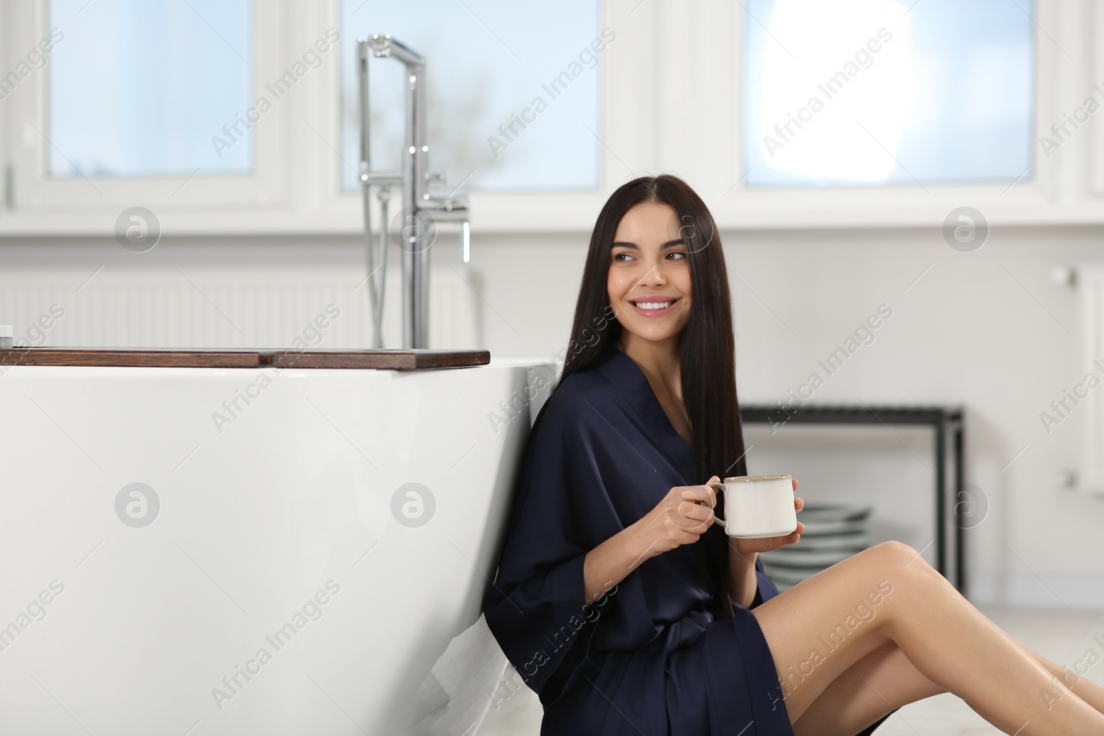 Photo of Beautiful happy woman in stylish bathrobe with cup of hot drink near tub in bathroom