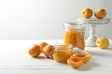 Photo of Jars of apricot jam and fresh fruits on white wooden table. Space for text
