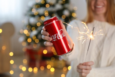 MYKOLAIV, UKRAINE - January 01, 2021: Woman with can of Coca-Cola and sparkler against blurred Christmas tree indoors, closeup