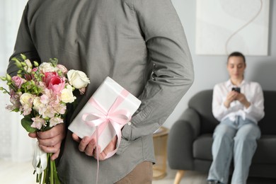 Photo of Man hiding bouquet of flowers and present for his beloved woman indoors, closeup