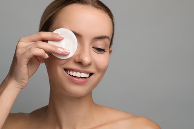 Photo of Smiling woman removing makeup with cotton pad on grey background, closeup. Space for text