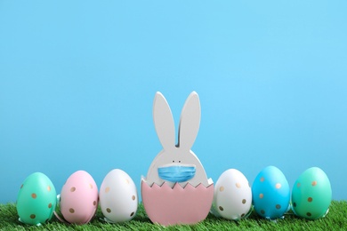 Image of COVID-19 pandemic. Easter bunny figure in protective mask and dyed eggs on green grass against light blue background