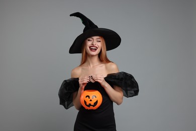 Photo of Happy young woman in scary witch costume with pumpkin bucket on light grey background. Halloween celebration