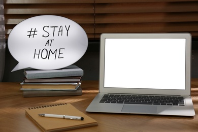 Photo of Laptop, office supplies and speech bubble with hashtag STAY AT HOME on wooden table indoors. Message to promote self-isolation during COVID‑19 pandemic