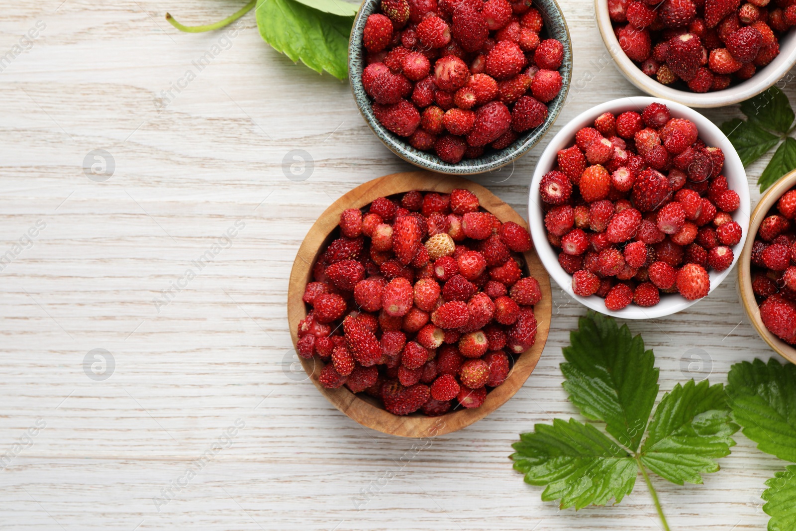 Photo of Fresh wild strawberries in bowls and leaves on white wooden table, flat lay. Space for text