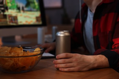 Photo of Young man with energy drink and chips playing video game at wooden desk indoors, closeup
