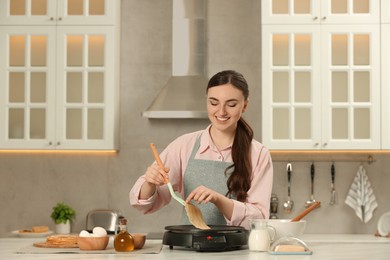 Photo of Happy woman cooking delicious crepe on electric maker at white marble table in kitchen