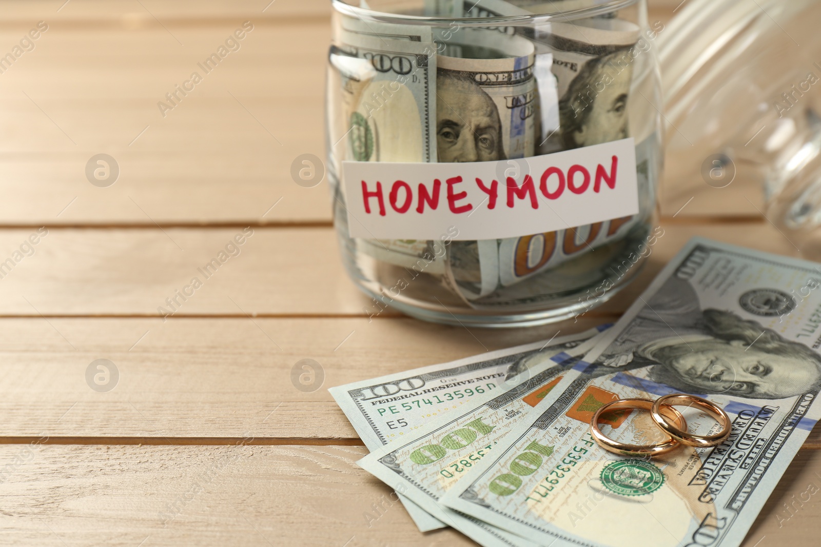 Photo of Glass jar with word Honeymoon, dollar banknotes and golden rings on wooden table, closeup