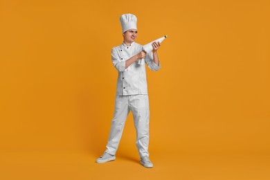 Photo of Portrait of happy confectioner in uniform holding piping bag on orange background