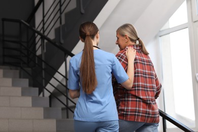 Photo of Young healthcare worker assisting senior woman on stairs indoors, low angle view
