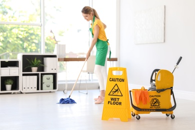 Photo of Phrase "CAUTION WET FLOOR" on safety sign, mop bucket with cleaning supplies and young woman on background