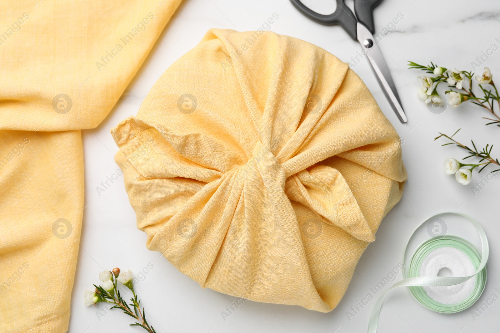 Photo of Furoshiki. Things packed in fabric, scissors, ribbon and flowers on white marble table, flat lay