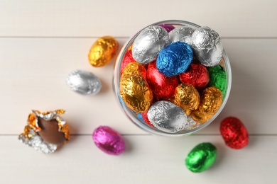 Glass jar with chocolate eggs wrapped in colorful foil on white wooden table, flat lay