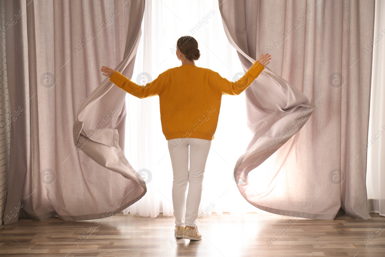 Photo of Woman opening window curtains at home in morning, back view