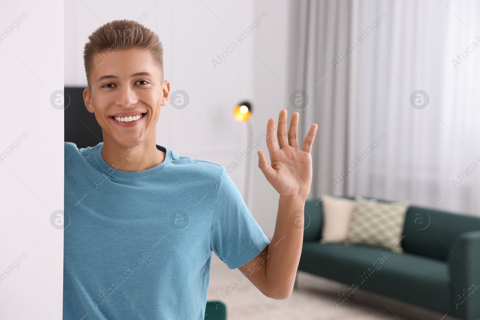 Photo of Happy man waving near white wall at home, space for text. Invitation to come in room