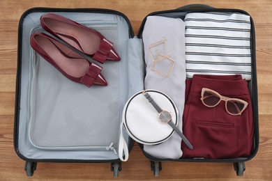 Open suitcase with female clothing, shoes and accessories on wooden background