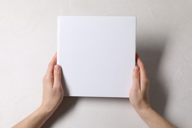 Woman holding book with blank cover at white table, top view. Mockup for design