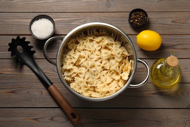 Cooked pasta in metal colander, lemon, oil and spices on wooden table, flat lay