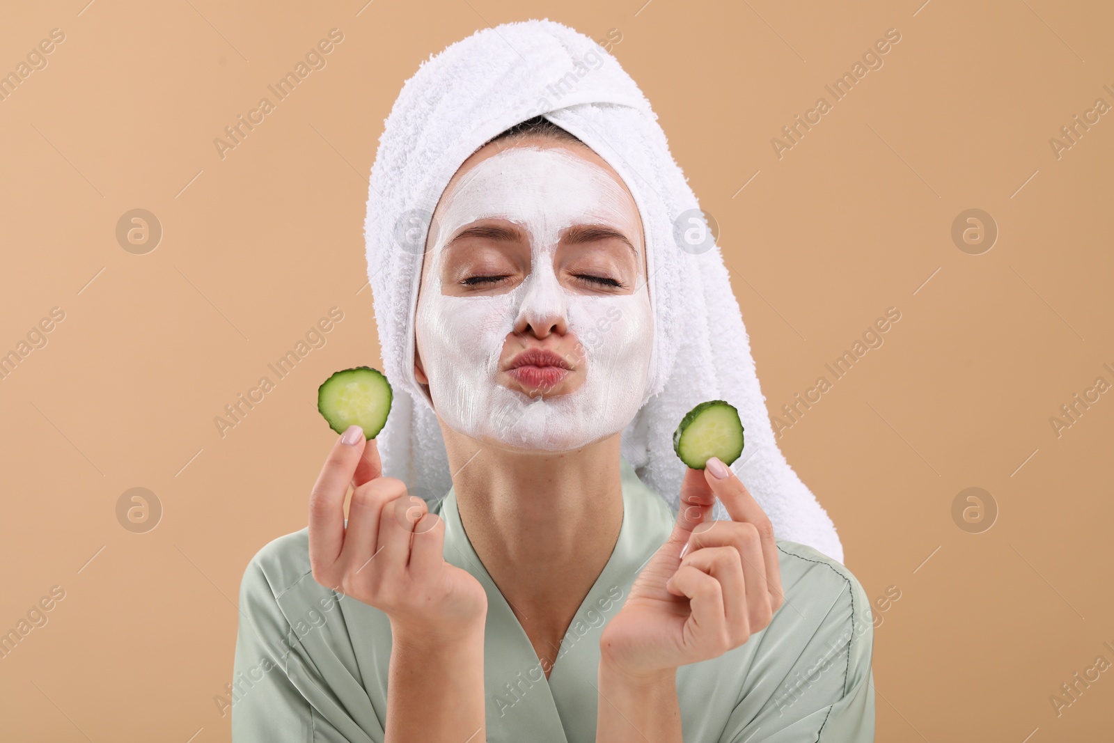 Photo of Woman with face mask and cucumber slices sending air kiss on beige background. Spa treatments