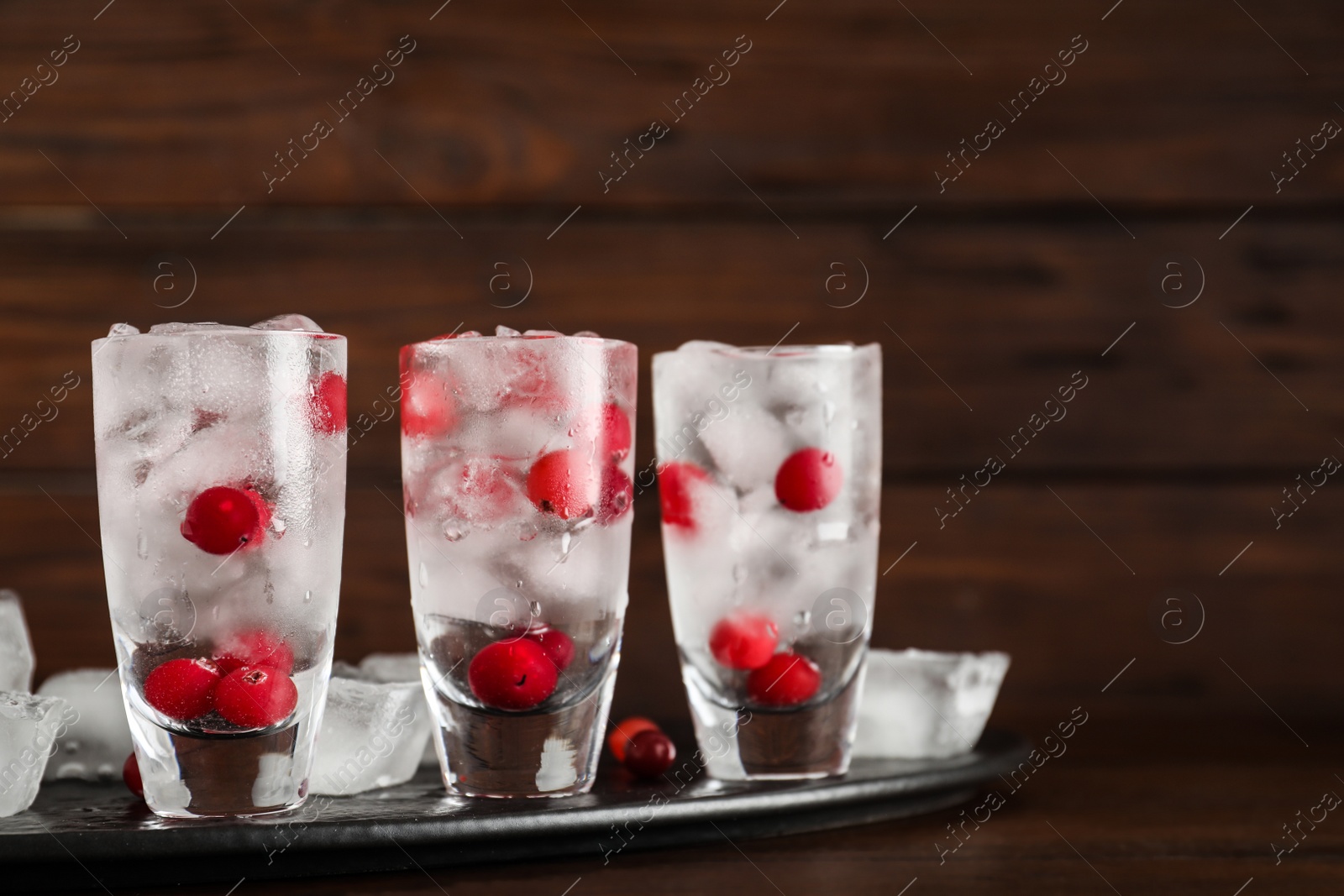 Photo of Shot glasses with vodka, ice and cranberries on table against wooden background