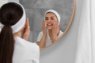 Photo of Young woman with headband washing her face near mirror in bathroom