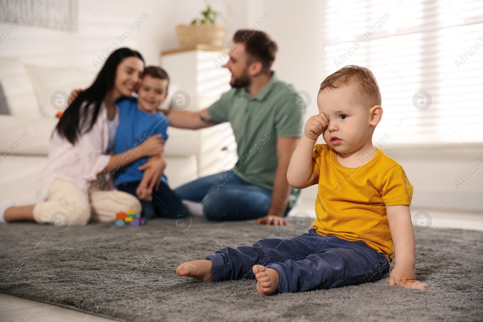 Photo of Unhappy baby sitting alone on floor while parents spending time with his elder brother at home. Jealousy in family