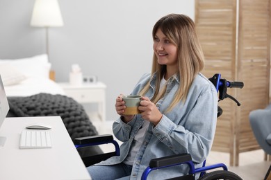 Woman in wheelchair with cup of drink using computer at home