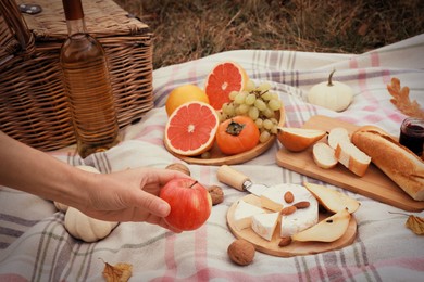 Image of Woman taking apple from blanket with snacks and wine outdoors, closeup. Autumn picnic