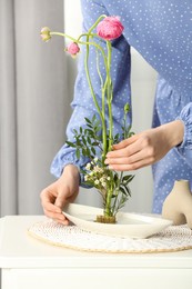 Photo of Woman creating stylish ikebana with beautiful flowers and green branches at white table, closeup