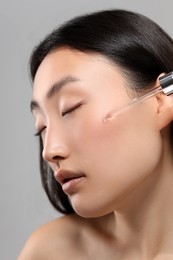 Beautiful young woman applying cosmetic serum onto her face on grey background, closeup