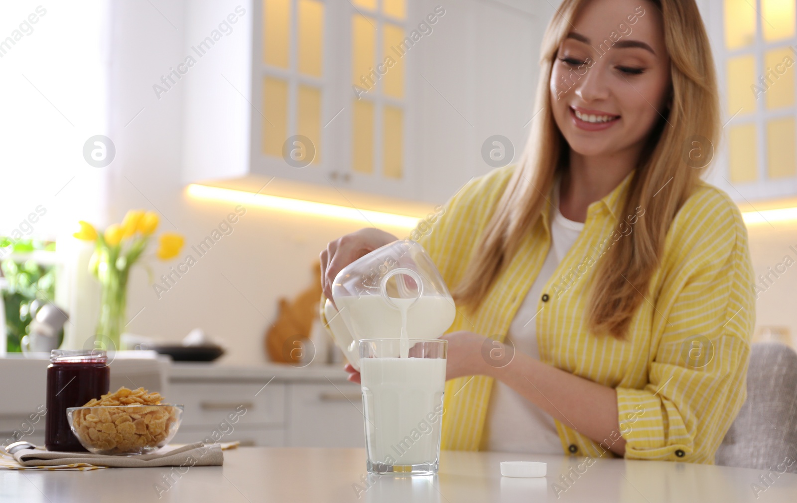 Photo of Young woman pouring milk from gallon bottle into glass at white table in kitchen