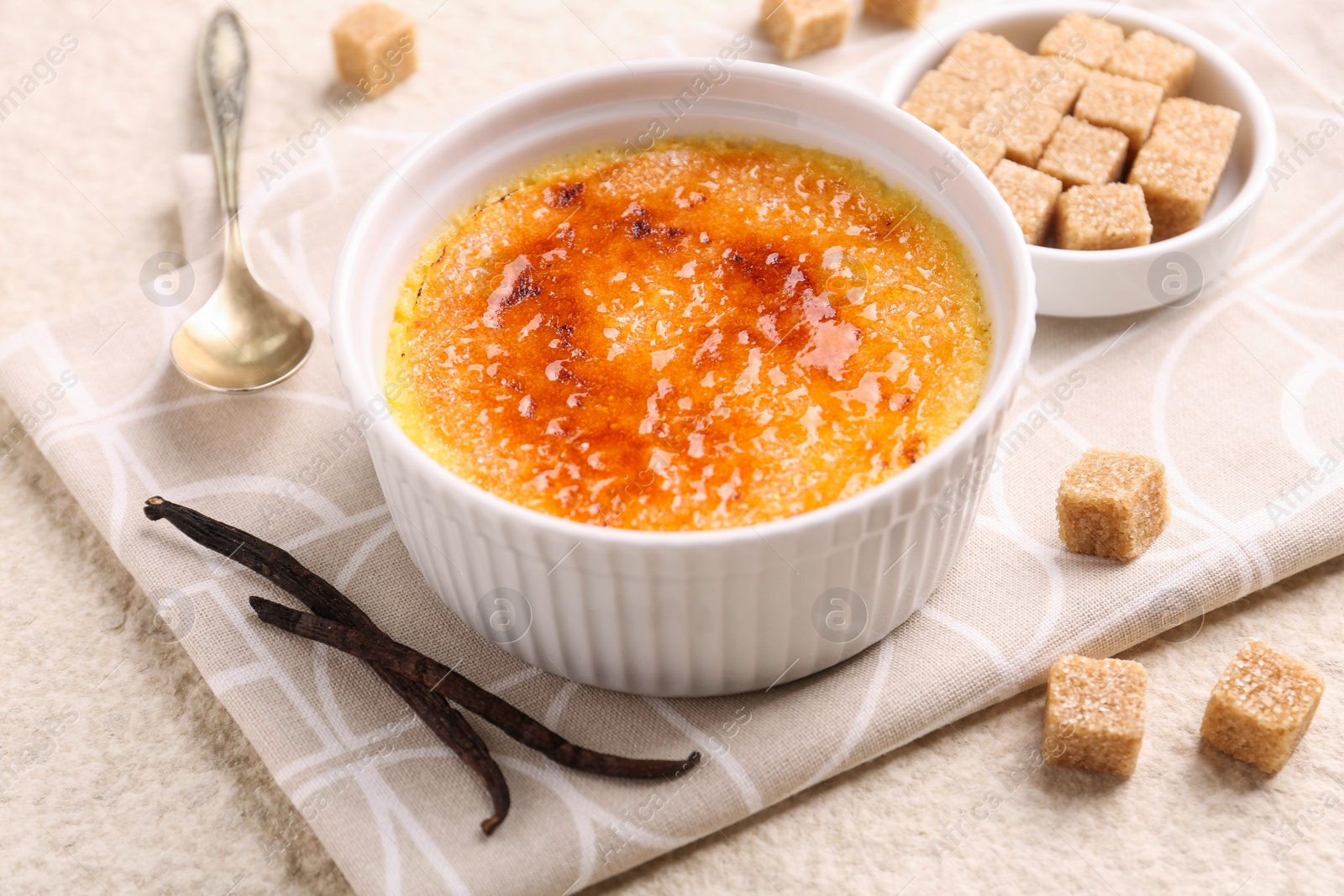 Photo of Delicious creme brulee in bowl, vanilla pods and sugar cubes on light textured table, closeup