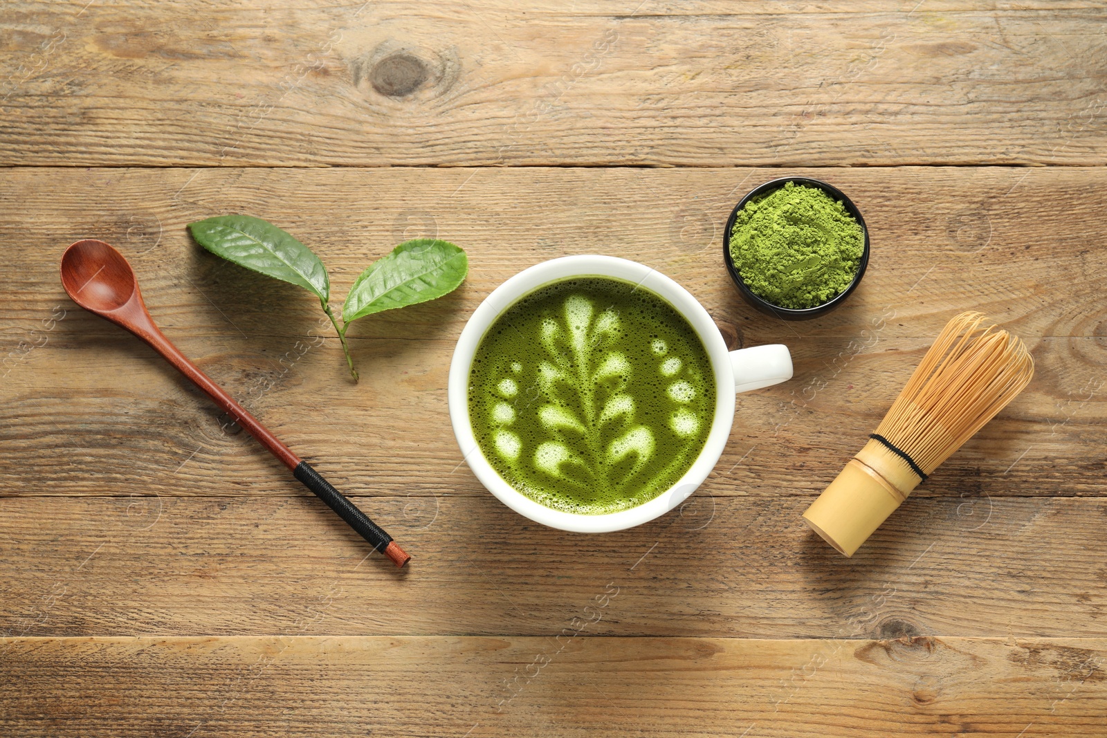 Photo of Delicious matcha latte, powder, spoon, leaves and whisk on wooden table, flat lay